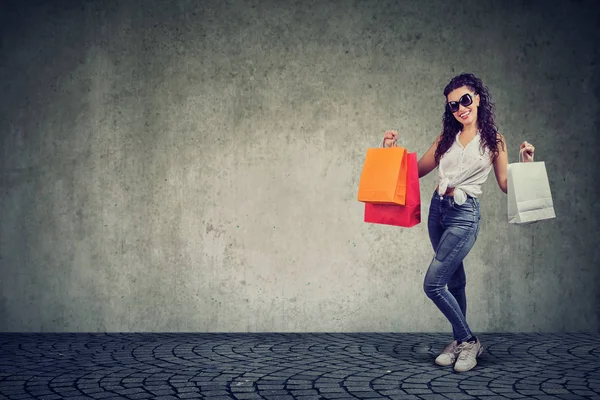 内容年轻的褐发女郎在太阳镜站在灰色背景五颜六色的购物袋 — 图库照片