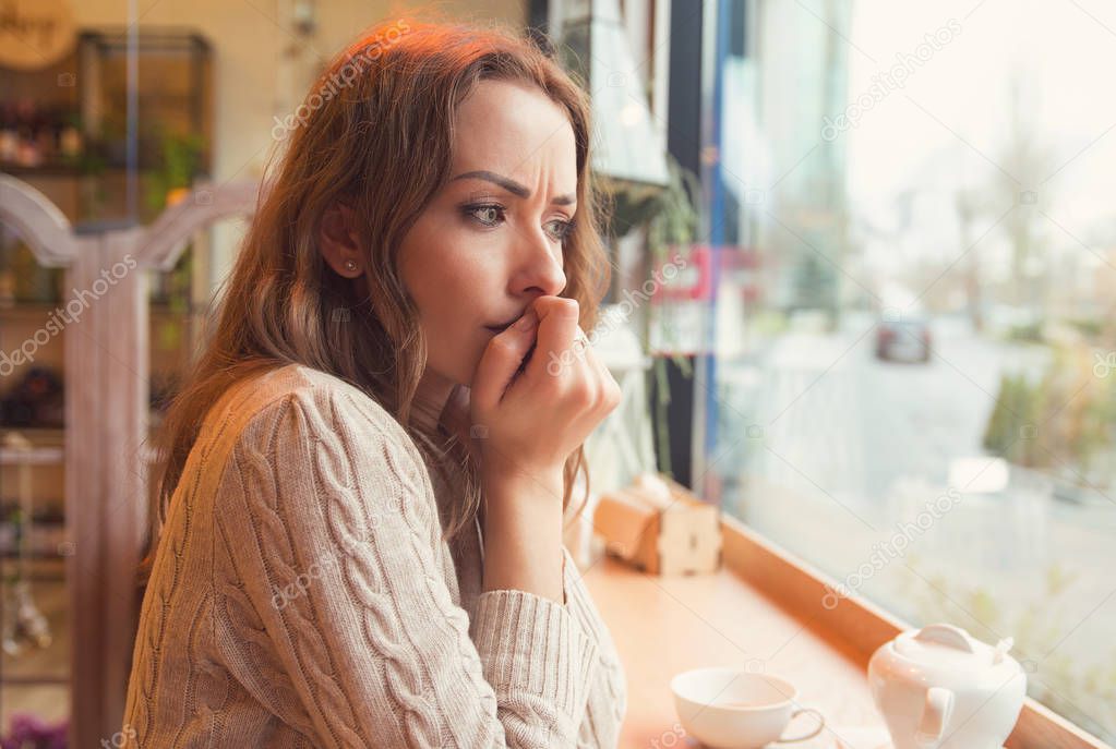 Nervous worried young woman biting nails and looking away sitting alone in a coffee shop