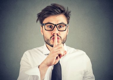 Sessizlik için sorma ve gri arka plan kameraya gülümseyen dudaklarında parmak tutan Gözlük Genç hipster adam