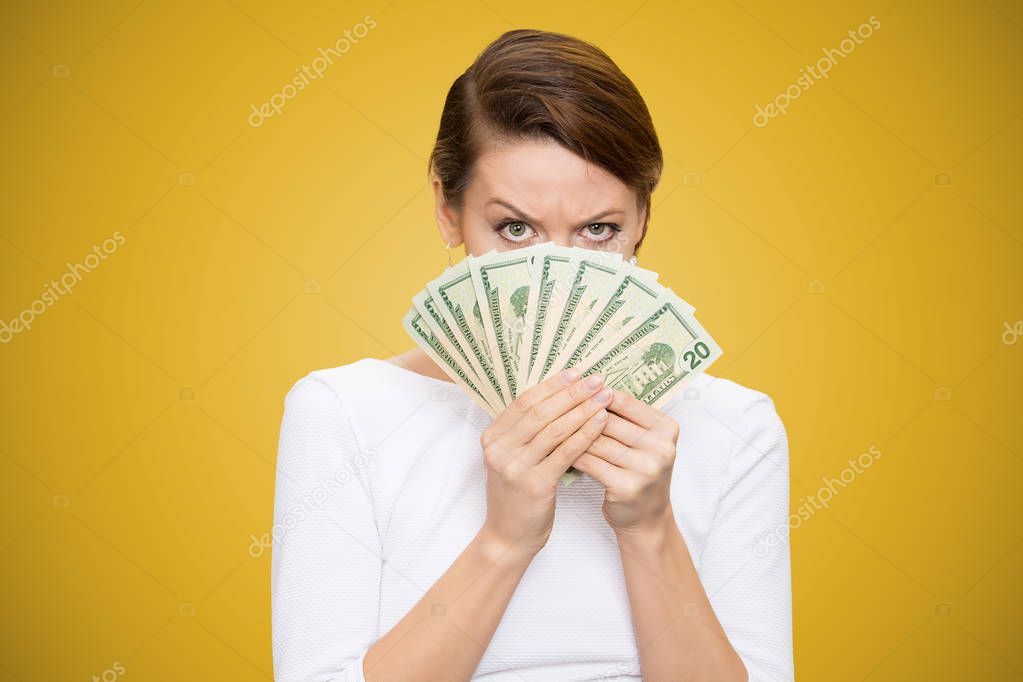 Young grumpy woman covering face with heap of bills looking at camera on yellow background