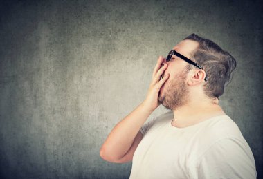 Tombul adam beyaz t-shirt ve gri arka plan üzerinde esneme sırasında ağız kapsayan gözlük yan görünüm