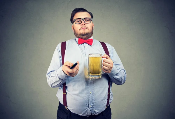 Adult lazy obese man in formal outfit holding mug of beer and watching TV on gray background