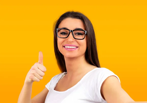 一个快乐美丽的女人的肖像显示竖起大拇指的手势 而采取在黄色背景隔离的自拍 — 图库照片