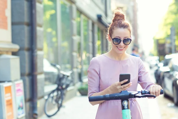 Donna felice in occhiali da sole utilizzando smartphone e noleggiare scooter elettrico moderno con un'applicazione — Foto Stock