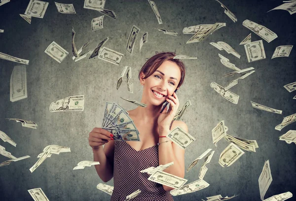 Attractive woman talking on a smartphone holding money, cashback under dollar rain — Stok fotoğraf