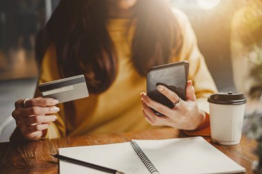 Genç, güzel Asyalı bir kadın, akıllı telefon ve kredi kartını kullanarak kafede online alışveriş yapıyor.