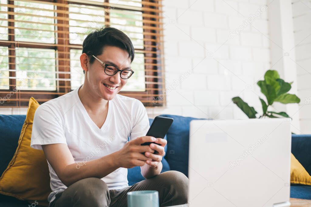 Young Asian man using phone for online shopping, and working with laptop at home