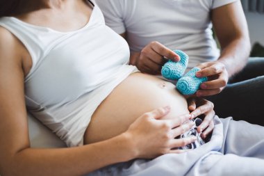 Genç Asyalı koca hamile karısıyla beyaz yatakta mutlu ve gülümseyerek oynuyor.