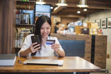 Genç, güzel Asyalı bir kadın kafeteryada online alışveriş yapmak için akıllı telefon ve kredi kartı kullanıyor.