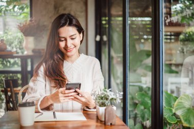 Genç Asyalı güzel bir kadın akıllı telefonu iş için kullanıyor, online alışveriş, para transferi, finans, internet bankacılığı. Kahve dükkanında.