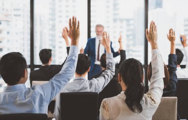 Bir grup iş adamı toplantı odasındaki seminerde soru sormak ve konuşmacılara cevap vermek için ellerini kaldırıyor.