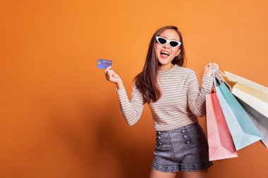 Güzel, modaya uygun bir Asyalı kadın elinde telefon ve kredi kartıyla turuncu arka planda çekim yapıyor.