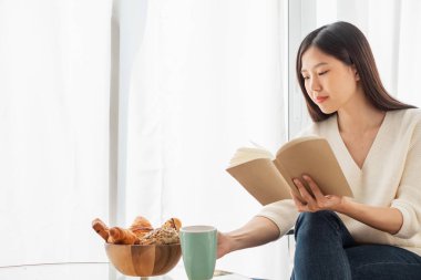 Genç ve güzel Asyalı kadın oturma odasında dinleniyor, kitap okuyor, sabahları mutlu bir şekilde kahve içiyor.