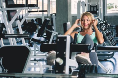 Spor salonunda oturup ağırlık antrenmanı yapan bir kadın. Mutlu ve gülümseyen, spor salonu konsepti olan.