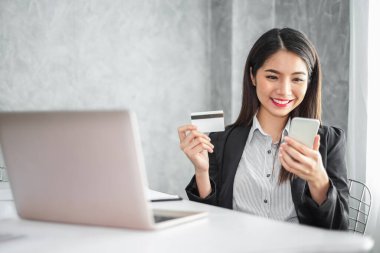 Takım elbiseli Asyalı genç iş kadını internet alışverişi, internet bankacılığı ve internet üzerinden satın alma hizmetleri için akıllı telefon ve kredi kartı kullanıyor..