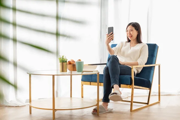 Young beautiful Asian woman relaxing in living room at home, reading books, drinking coffee and using phone in the morning with happy feeling