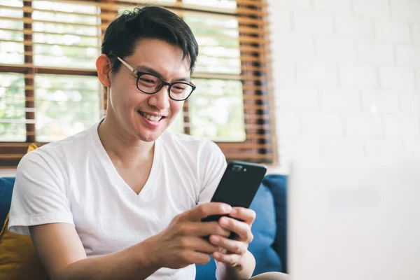 Genç Asyalı adam online alışveriş için telefon kullanıyor ve evde dizüstü bilgisayarla çalışıyor.