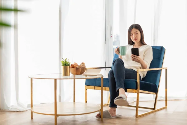 自宅のリビングルームでリラックスした若いアジアの女性 読書本 コーヒーを飲み 幸せな気持ちで午前中に携帯電話を使用して — ストック写真