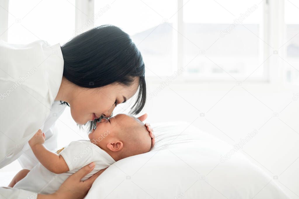 Young Asian mother kissing her newborn baby with love on white bed, mother's day concept