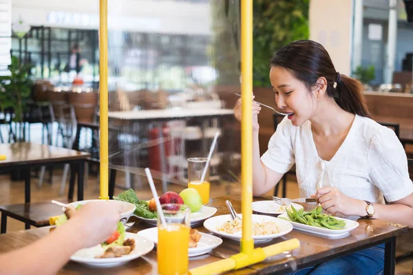 Asian woman sitting separated in restaurant eating food with table shield plastic partition to protect infection from coronavirus covid-19, restaurant and social distancing concept