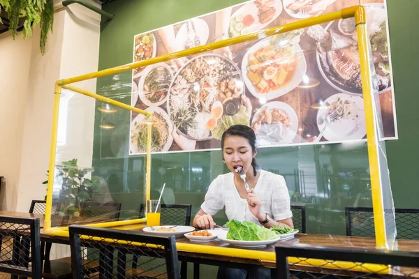 Asian woman sitting separated in restaurant eating food with table shield plastic partition to protect infection from coronavirus covid-19, restaurant and social distancing concept