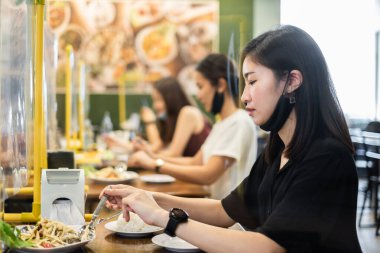 Restoranda ayrı oturan Asyalı erkek ve kadın Coronavirus covid-19 enfeksiyonunu, yeni normal restoran ve sosyal uzaklık kavramını korumak için masa kaplamalı plastik bölmeli yiyecekler yiyorlar.