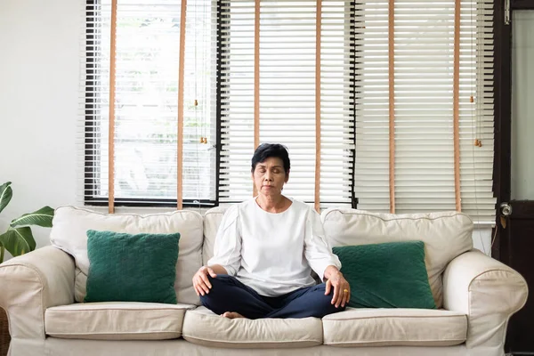 Senior old Asian woman sitting on sofa praticing yoga and meditation at home, retirement and wellbeing concept