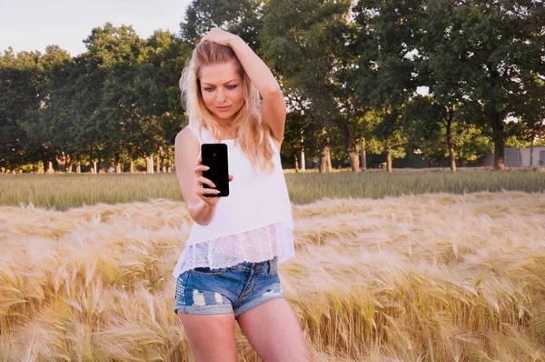 A beautiful girl at sunrise in a field of wheat makes selfie. The girl is glad that summer has come and is photographing herself. A young girl walks on the wheat field at dawn and rejoices in the warm