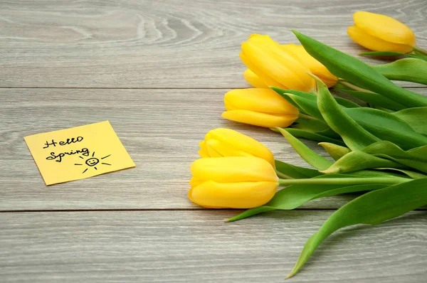 Beautiful bouquet of yellow tulips lies on a wooden gray table close up. Delicate flowers on the table with a note on the sticker hello spring.