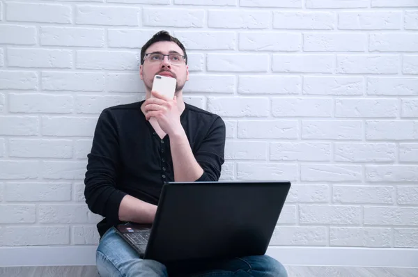 A young guy sits thoughtfully with a computer while holding a phone in his hand against the background of a white loft. The guy works at a computer at home. The guy working as a freelancer.