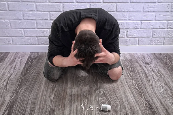 A young guy is trying to commit suicide. The guy is trying to give up drugs. The guy is sitting on the floor with scattered pills on a white background.