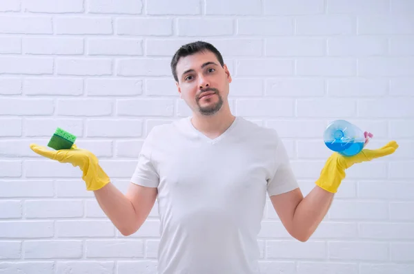 A young guy holds a sponge and detergent in his hands against a white loft. A guy in yellow, rubber gloves cleans the house.