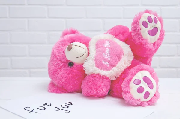 A pink toy bear lies on a table against the background of a white loft. Toy bear with a note on the table close-up. Holiday Greeting Card. Holiday background.