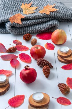 Küçük mumlar, koniler, nar, elmalar, kuru kırmızı yapraklar, beyaz ahşap bir masaya örülmüş gri eşarp. Merhaba sonbahar. Samimi sonbahar arkaplanı.