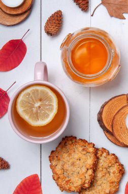 Kavanozda limon ve bal, yulaf ezmeli kurabiyeler, koniler, kuru yapraklar ve beyaz ahşap masa arka planında küçük mumlar olan bir fincan çay. Sağlıklı kahvaltı. Samimi sonbahar arkaplanı.