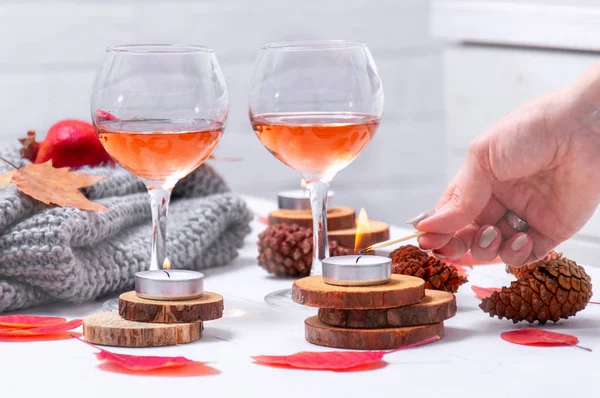 Small burning candles, two glasses with rose wine, cones, dry red leaves, gray scarf knitted on a white wooden table. Girl lights candles on the table. Romantic evening. Hello, Autumn. Cozy autumn bac