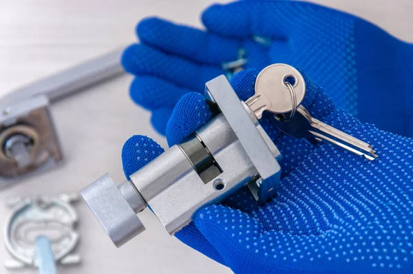 The guy holds in his hands in blue gloves the core of the lock and door keys on a light background. Repair in the house. Installing a new lock on the door.