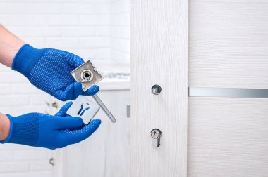 Adam yeni kapı kolları takıyor ve odanın kapısını kilitliyor. Evde tamirat var. Mavi eldivenli adam kapı kollarını tutuyor. Kapı kilidi takılıyor.