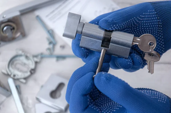 Adam elinde mavi eldivenlerle, arka planda kilit ve kapı anahtarlarını tutuyor. Evde tamirat var. Kapıya yeni bir kilit takılıyor.. 