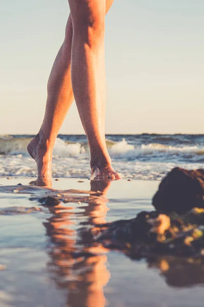 Young woman walking barefoot on sand beach. Close up view to female feet. Reflection in the water. Vertical view