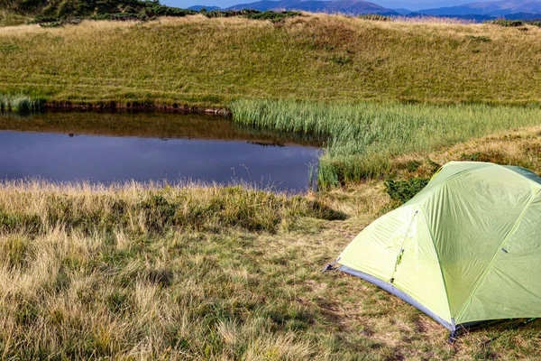 Camping tent on the shore of a mountain pond, surrounded by nature and hills. Outdoor travel and adventure concept. Green tent stands by a calm pond in the mountains, reflecting the beauty of wild nature and peaceful outdoor life.