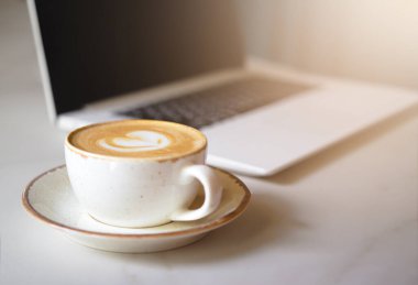 Kafedeki masanın üstündeki köpüklü kalp şeklinde beyaz kahve fincanını kapatır mısın? Bir fincan tabağında kapuçino. Arka planda bir dizüstü bilgisayar