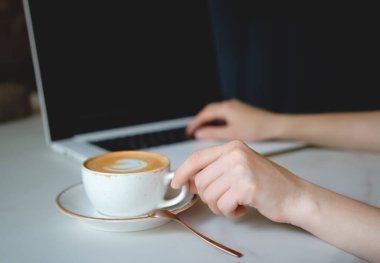 Bir kadın eli yakın planda beyaz bir latte fincanı alır. Kahve bir fincan tabağının üzerinde duruyor. Dizüstü bilgisayarda el izi mesajı. Kapuçino. Eline yavaşça odaklan. Dizüstü bilgisayarı kullanan adam.