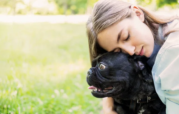Mooie jonge vrouw knuffelt een haar kleine Franse bulldog in een park buiten. Close-up portret van tevreden meisje met lang bruin haar omarmen grappige hond met ogen dicht. — Stockfoto
