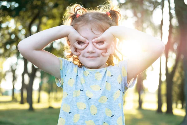 A beautiful little girl in the park fools around in the sunset. Elementary age girl is making face, smiling while looking at the camera. She has her hands near her cheeks makes glasses