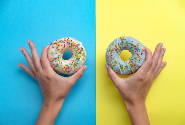 Beyaz ve mavi kremalı iki çörek. Renkli şekerli donutların genel manzarası. Sarı ve mavi arka planda donutları tutan kadın eller. Üst Manzara. Tatlı, diyet konsepti.