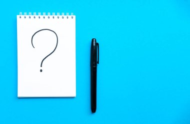 Defter ve kalemle en baştan çekim programı. Spiral defter mavi arka plan. Arkaplanda not defteriyle yazılmış soru işareti. Kopya uzayı ile yatay bileşim.