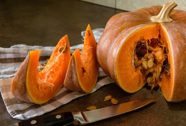 Balkabağıyla hayat devam ediyor. Dilimlenmiş bir balkabağı, iki dilim balkabağı, bir bıçak ve mutfak havlusu var..