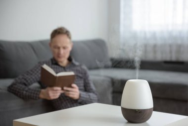 Arkaplanda beyaz bir masa üzerinde Aromo difüzör. Arka planda, bir adam kitap okuyor..