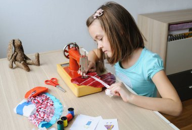 Kız dikiş makinesinde bir bebek için elbise dikiyor. Ürünü ölçmek için bir mezura kullanıyor..